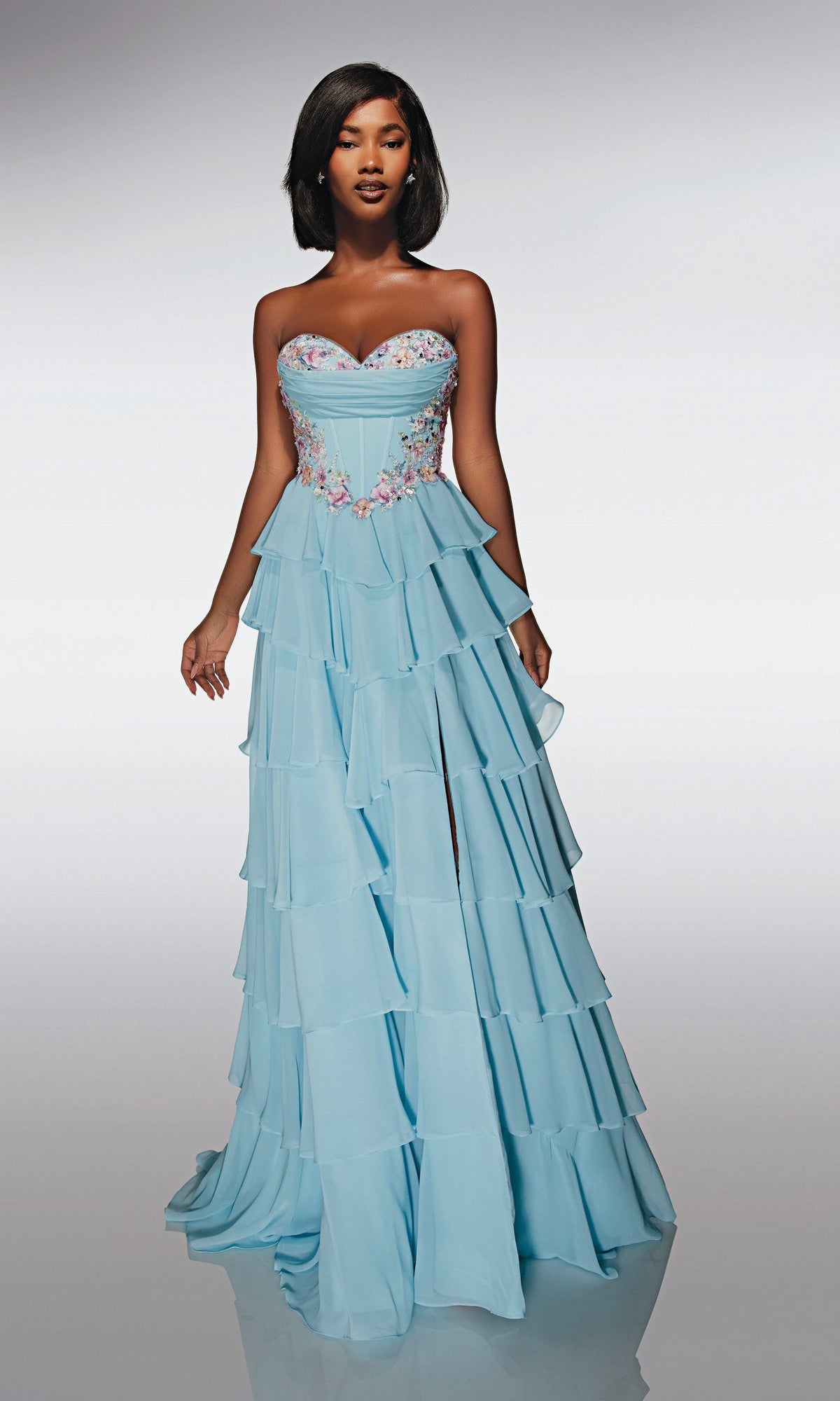 A woman stands against a plain background wearing the Alyce 62013 long prom dress, which is strapless chiffon with tiered ruffles and delicate floral appliques on the bodice.